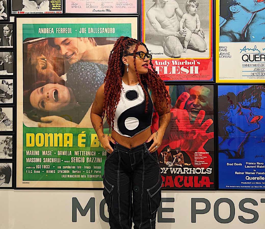 Donna Senders FunX met rode vlechten en yin-yang crop top poseert voor een kleurrijke muur met film- en kunstposters, symbool voor zelfexpressie en identiteit