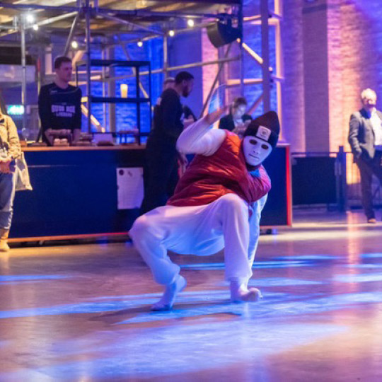 Randy Telg in een wit masker en rode outfit voert een krachtige move uit op een verlichte dansvloer, symbool voor zelfexpressie en performance