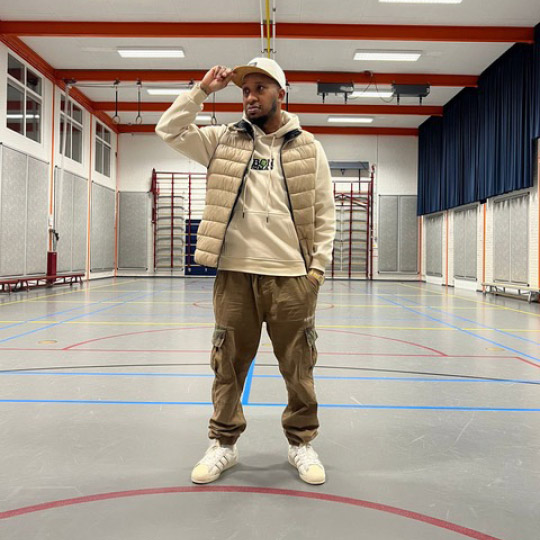 Randy Telg in beige streetwear-outfit staat in een gymzaal, symbool voor groei, zelfvertrouwen en roots