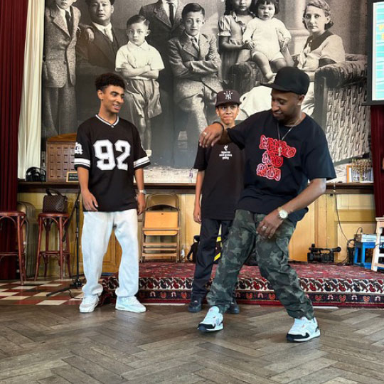 Drie jonge mannen dansen en lachen in een historische zaal, symbool voor community, cultuur en het delen van kennis